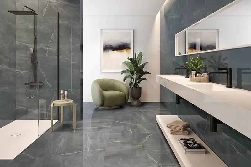 Full grey bathroom with a walk in shower on the left and green chair. Full length sink and mirror on the right.